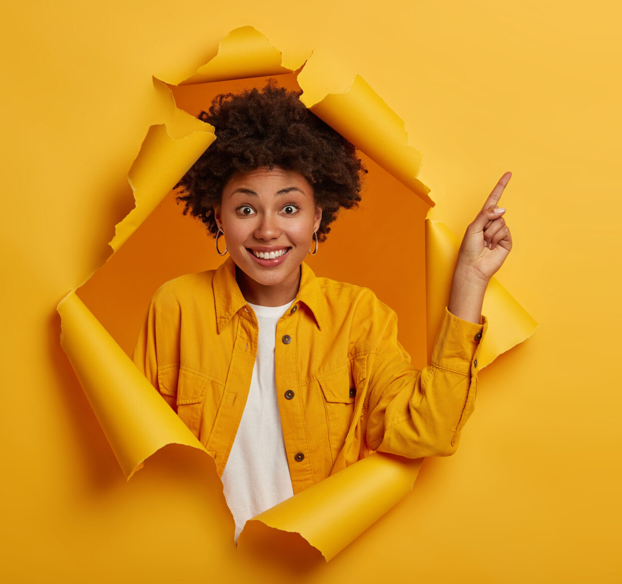Studio shot of positive African American woman points finger to copy space above, excited by good information, smiles pleasantly, wears yellow jacket, stands in ripped paper hole. Advertisement